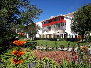 A two-story building with a red balcony, surrounded by a well-maintained garden with various plants and flowers. - 1BR Flat with terrace, parking and wifi in Lamalou (Lamalou-les-Bains)