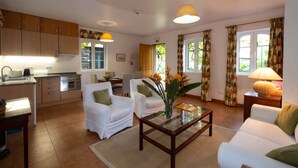 A modern kitchen with wooden cabinets, a dining area with white chairs, and a living room with a glass coffee table and white sofas. - Casa das Vinhas (Funchal)