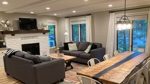 Una sala de estar moderna con chimenea, un sofá, una mesa de comedor con sillas y ventanas grandes con cortinas.