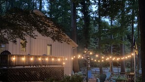 Una cabaña rústica con terraza de madera y luces cálidas, rodeada de árboles y mobiliario exterior.