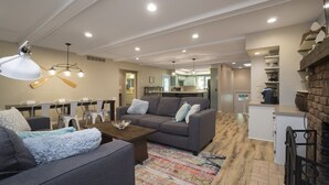 Amplia sala de estar con un sofá gris, una mesa de centro de madera y un tapiz colorido. Se observa un comedor con sillas blancas y un candelabro. La cocina se ve al fondo, con electrodomésticos modernos.