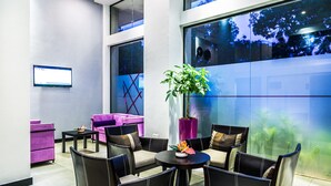 A modern lounge area with a round table, comfortable seating, a large window with a view of trees, and a mounted screen on the wall. - Home Residence Hotel (Cotonou)