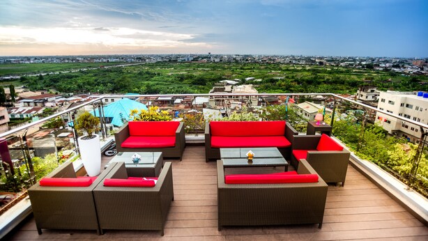 A rooftop terrace with wicker furniture and red cushions, offering a view of a cityscape and greenery. - Home Residence Hotel (Cotonou)