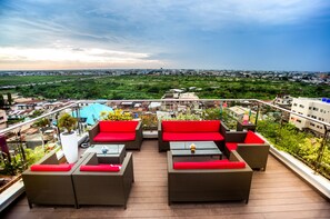 Terraza en la azotea con muebles de mimbre y cojines rojos, con vistas a un paisaje urbano y zonas verdes.
