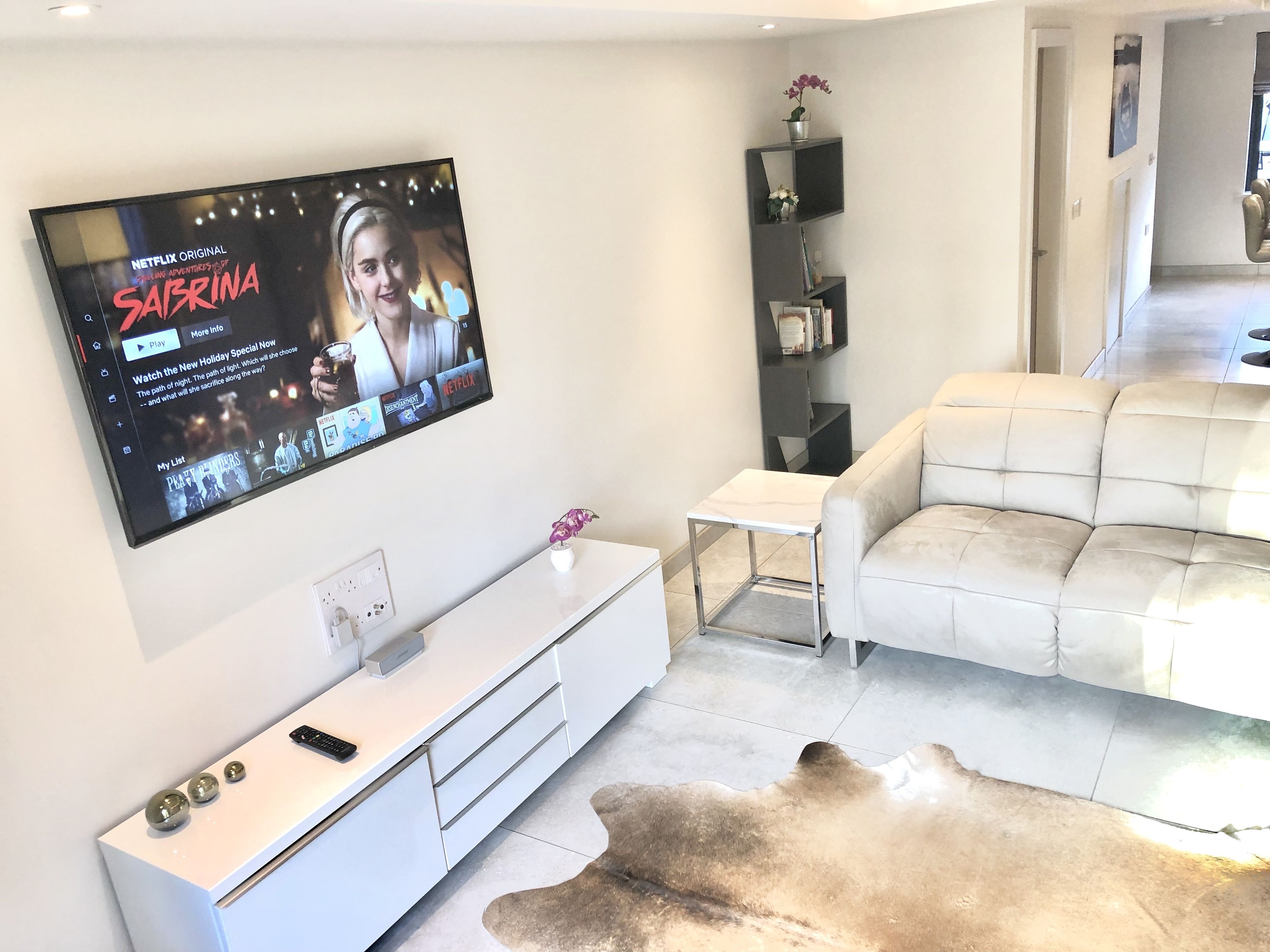 Un salon moderne avec un téléviseur à écran plat fixé au mur, un meuble de rangement blanc, un canapé beige et une petite table d’appoint blanche.