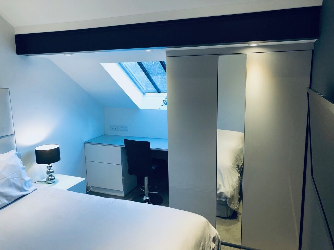 A bedroom with a bed, a desk, and a wardrobe. There is a skylight above the desk.