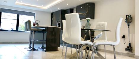A modern kitchen with a glass dining table, white chairs, and stainless steel appliances.