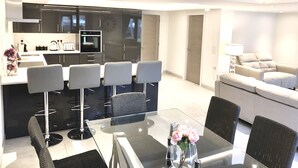 A modern kitchen with dark cabinets, a central island, and bar stools. There is a dining table with chairs and a vase of flowers. A sofa and a lamp are visible in the living area. - Millstream Mews (Bakewell)