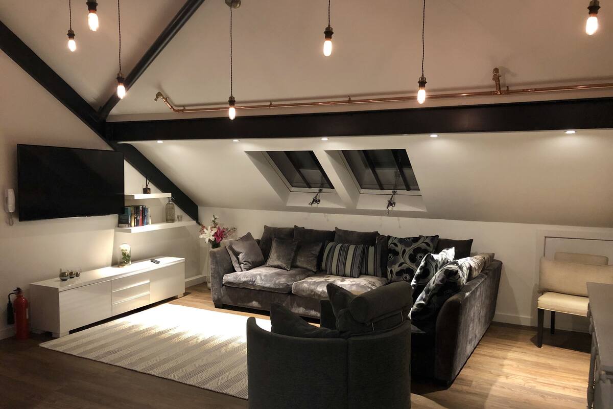 A modern loft apartment with a high ceiling, exposed beams, a large sectional sofa, a white console table, and a small side table.