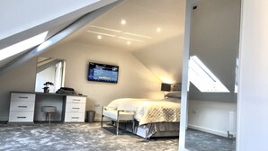 A bedroom with a sloped ceiling, a bed, a desk, and a TV mounted on the wall. - Millstream Mews (Bakewell)