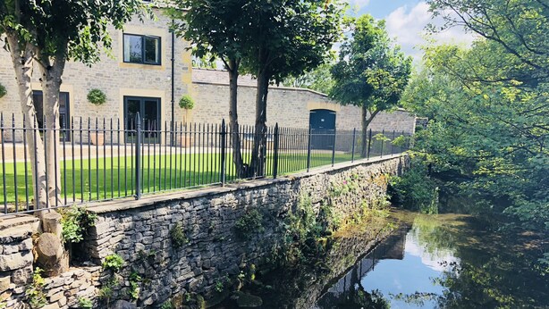 A stone bridge over a calm river, surrounded by lush greenery and a stone building with a black door. - Millstream Mews (Bakewell)