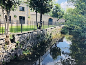 A stone bridge over a calm river, surrounded by lush greenery and a stone building with a black door.