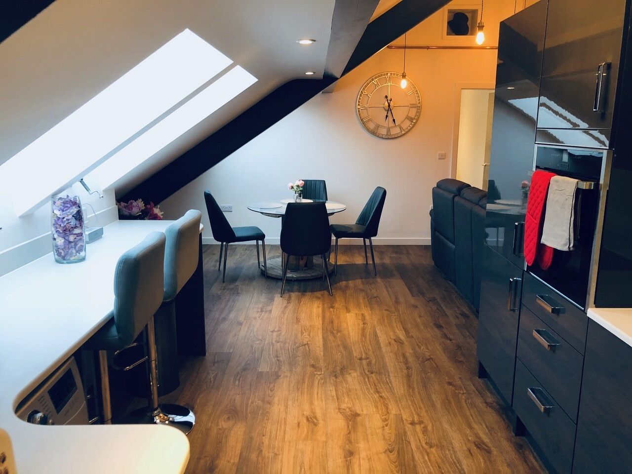 A modern kitchen with a skylight, wooden flooring, a dining area with chairs, and a black cabinet with a red towel.