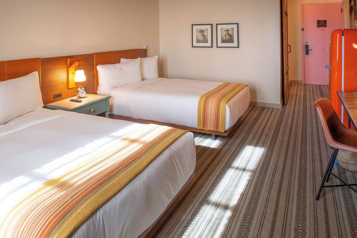 A hotel room with two beds, a nightstand with a lamp, a desk, and a chair.