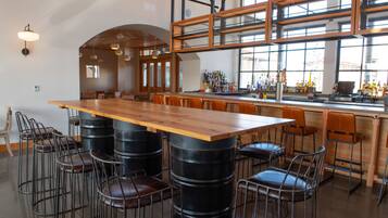 Un grand espace bar avec une longue table en bois, entourée de tabourets de bar en métal, de barils noirs et d’une variété de bouteilles exposées sur le comptoir.