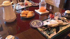Una mesa de comedor dispuesta con platos, una jarrón, una botella de zumo de naranja y un bol de fruta.