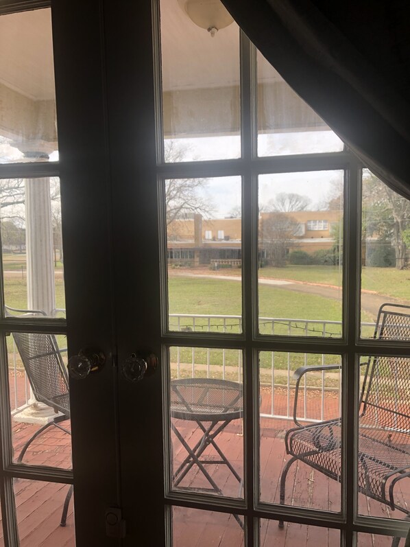 Vista desde una habitación con un ventanal grande que da a un patio con sillas de metal y una mesa.