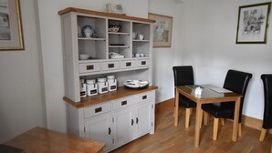 A dining area with a wooden table and chairs, a white cabinet with drawers, and a wall-mounted shelf with dishes and jars. - Gordon House (Wimborne)
