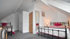 A bedroom with a sloped ceiling, a bed with a white and red bedspread, a bedside table with a lamp, and a door leading to another room. - Gordon House (Wimborne)
