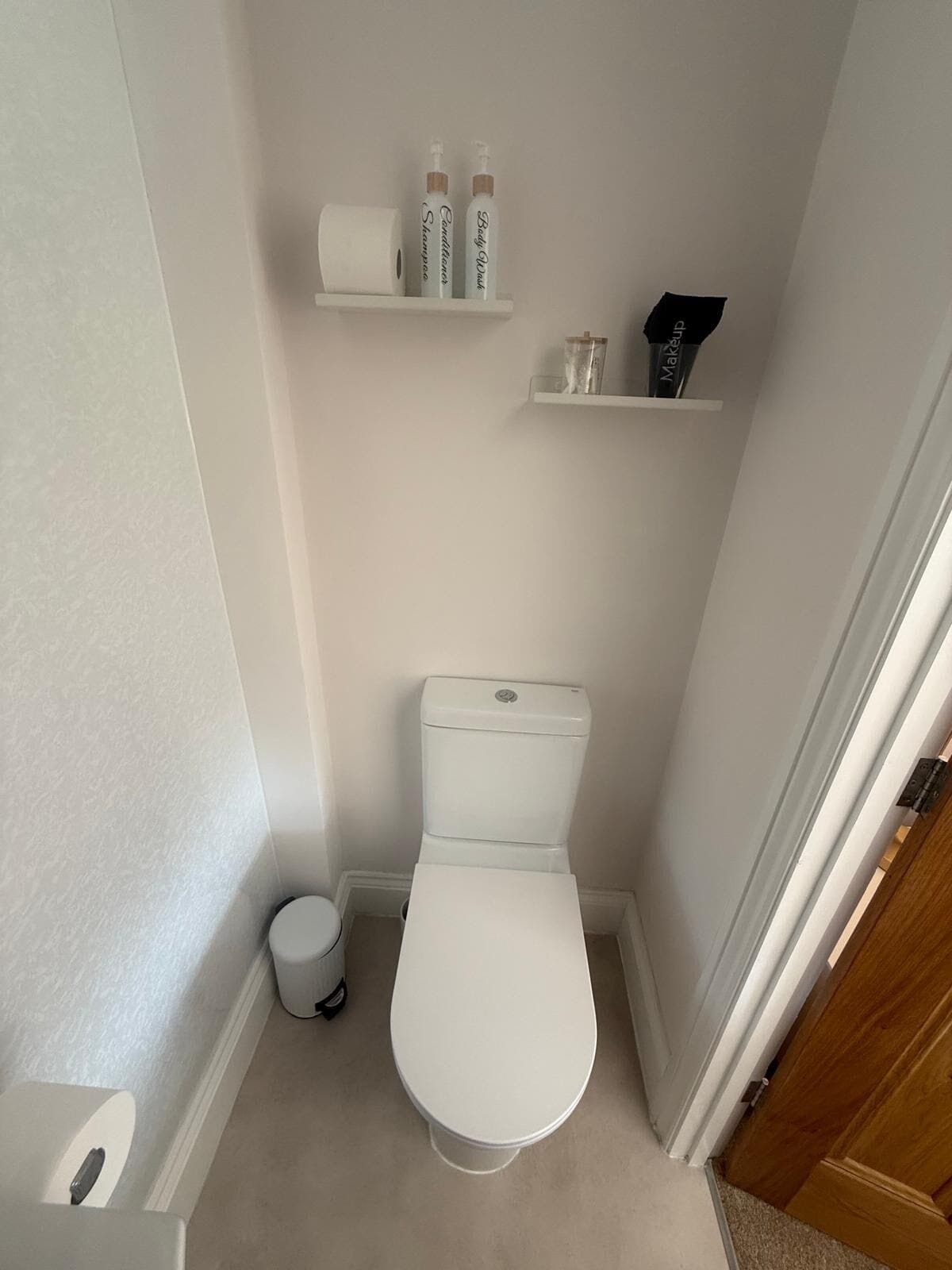 A small, white bathroom with a toilet, a roll of toilet paper, and two bottles of body wash on a shelf.