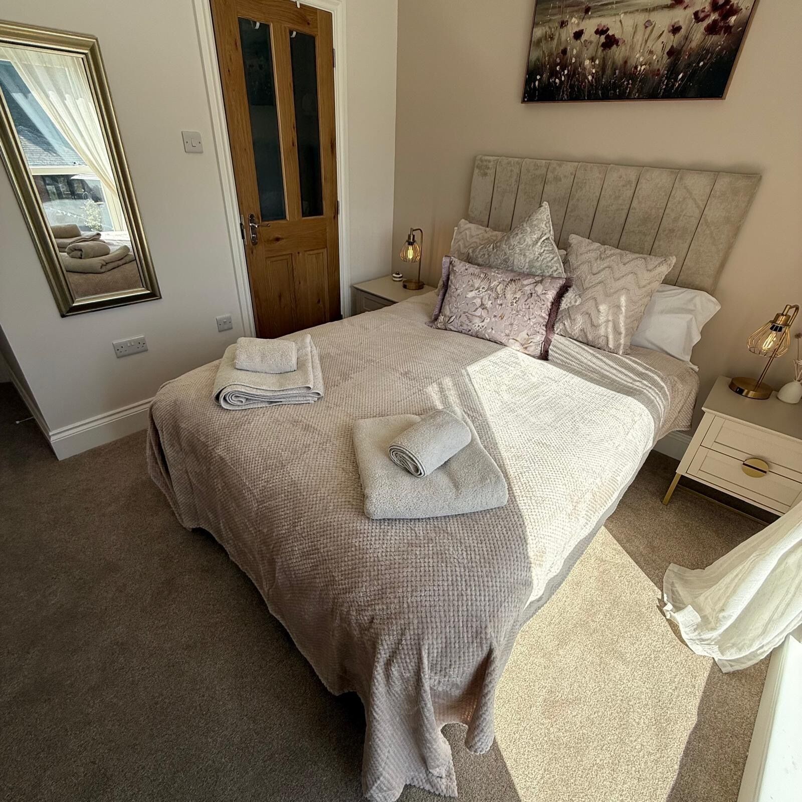 A bedroom with a bed, pillows, a nightstand, a lamp, a mirror, and a painting on the wall.