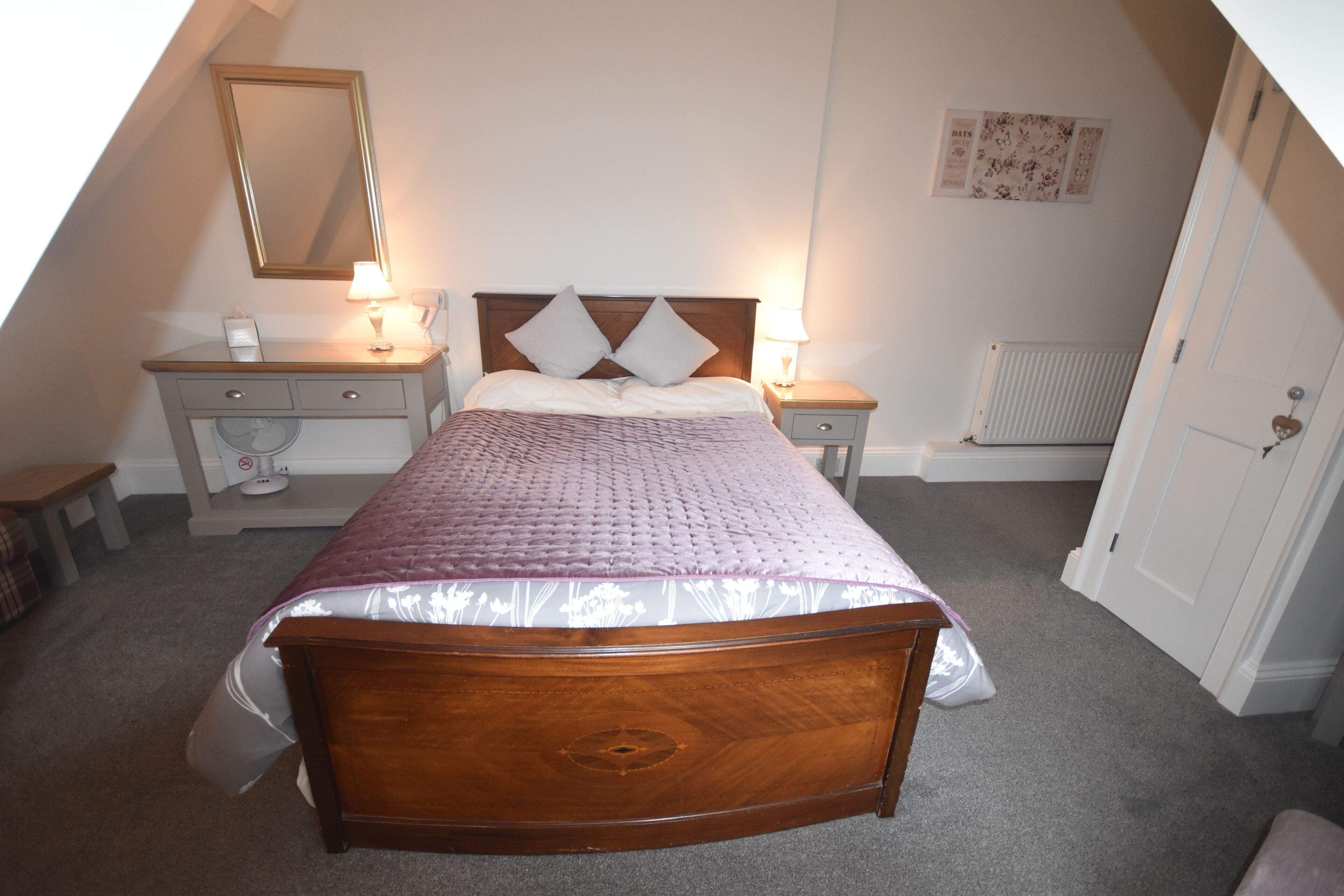 A bedroom with a wooden bed, two bedside tables with lamps, a radiator, and a door with a heart-shaped decoration.