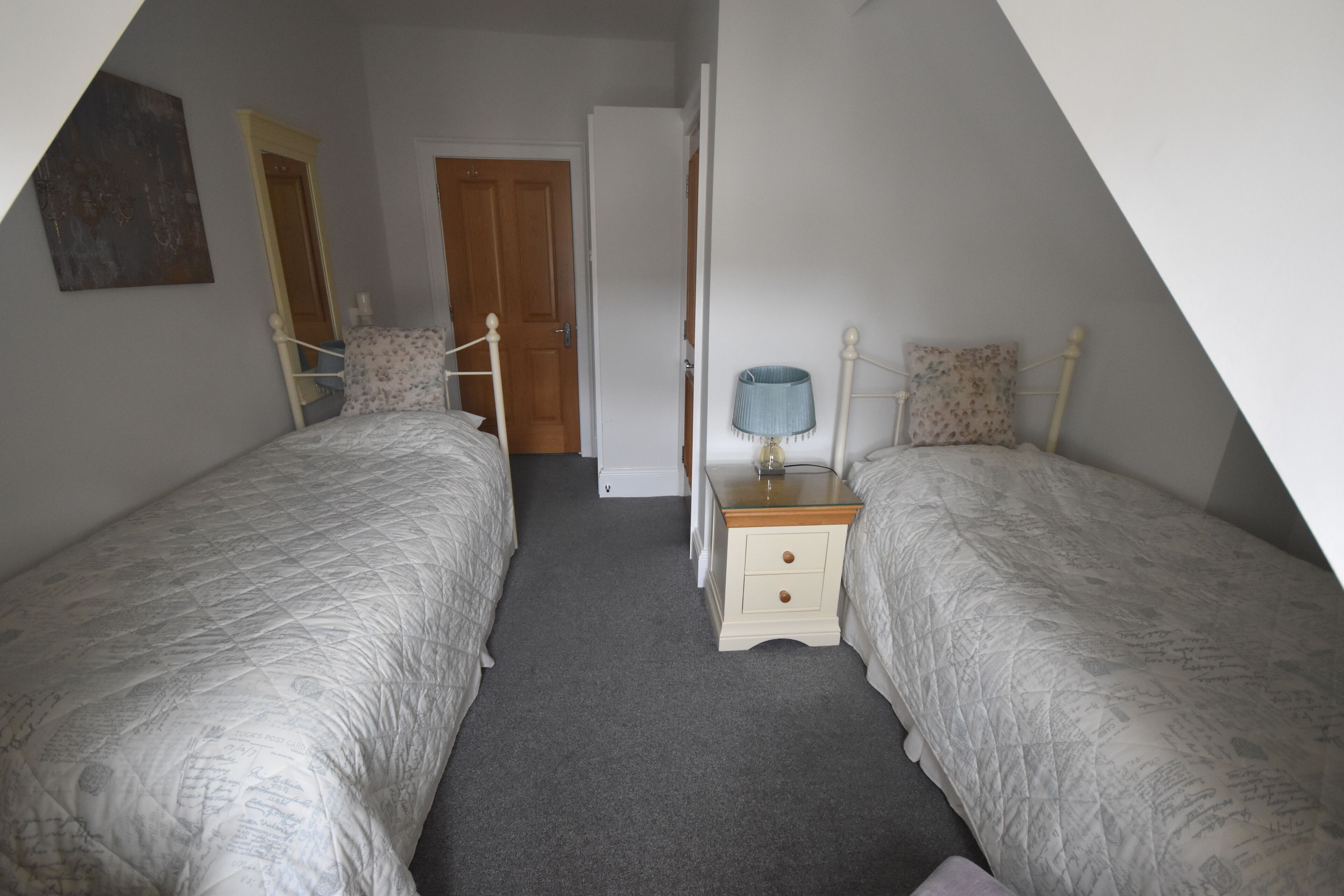 A bedroom with two single beds, a nightstand with a lamp, a door, and a wall-mounted artwork.