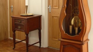 A room with a wooden floor, a tall grandfather clock, a wooden door, and a small wooden table with a decorative item on it. - Gordon House (Wimborne)