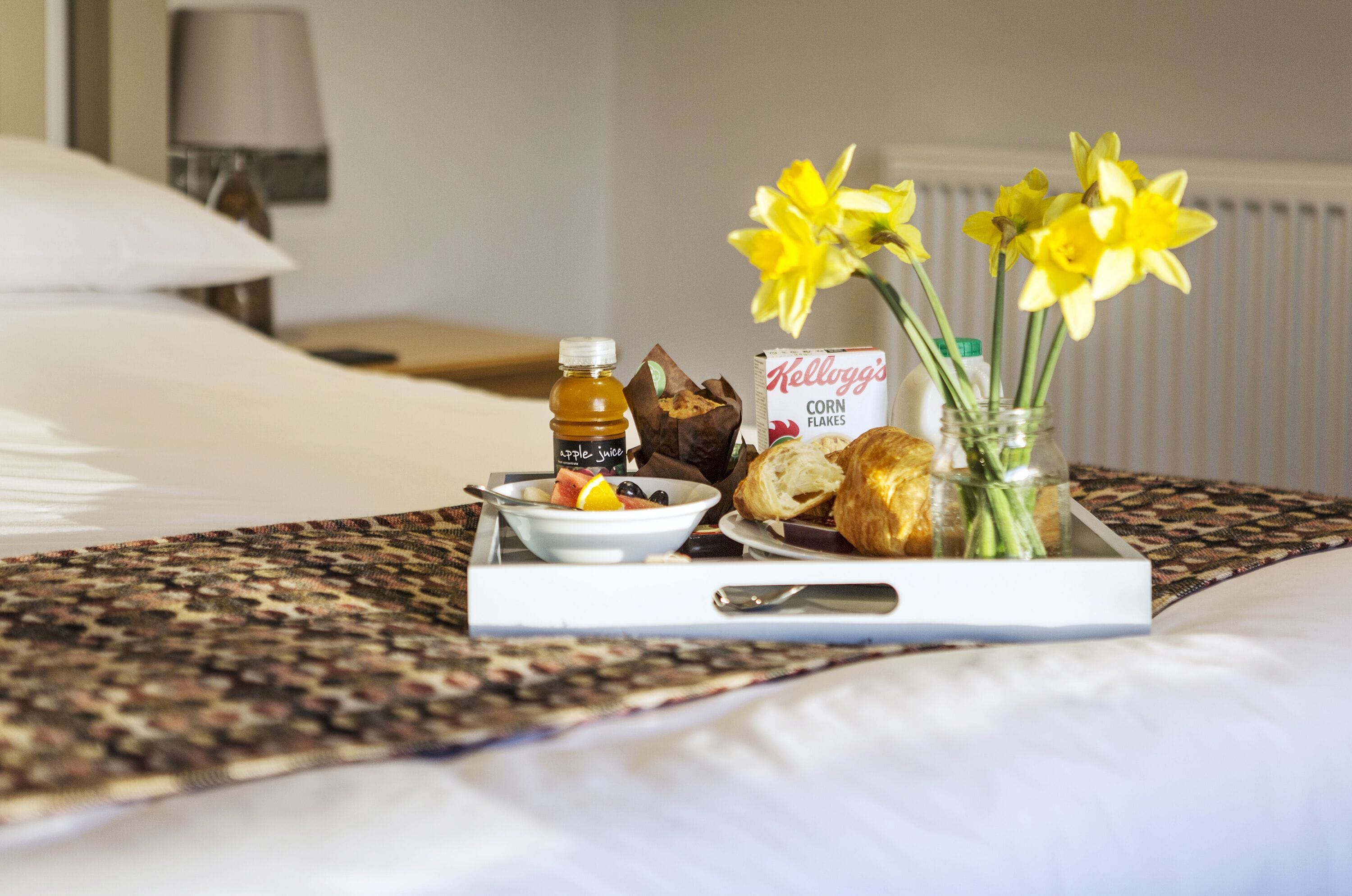 A breakfast tray with fruits, bread, and a bottle of juice on a bed with a patterned bedspread.