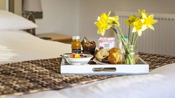 A breakfast tray with fruits, bread, and a bottle of juice on a bed with a patterned bedspread.