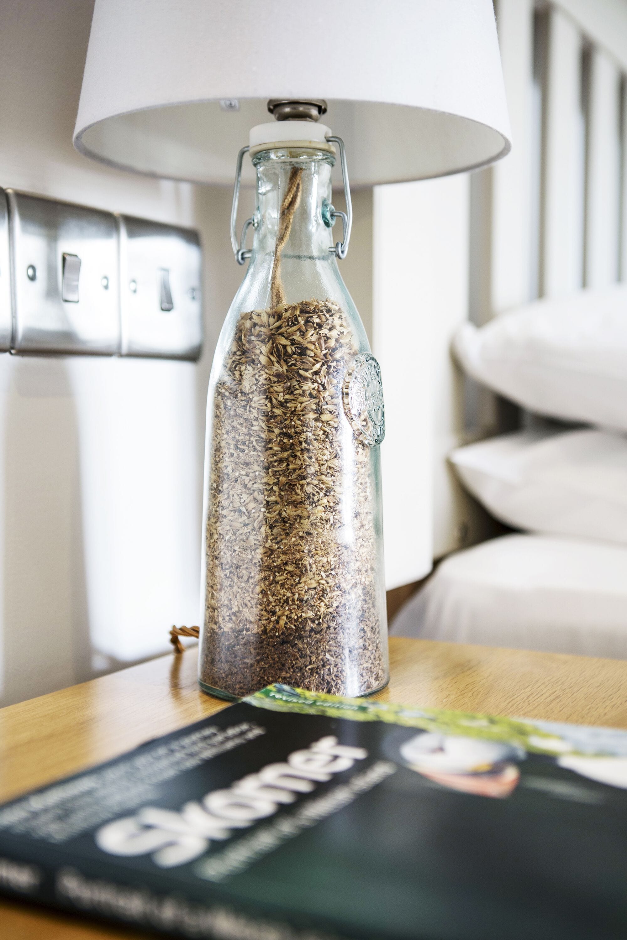 A clear glass bottle filled with seeds, placed on a wooden table with a magazine.