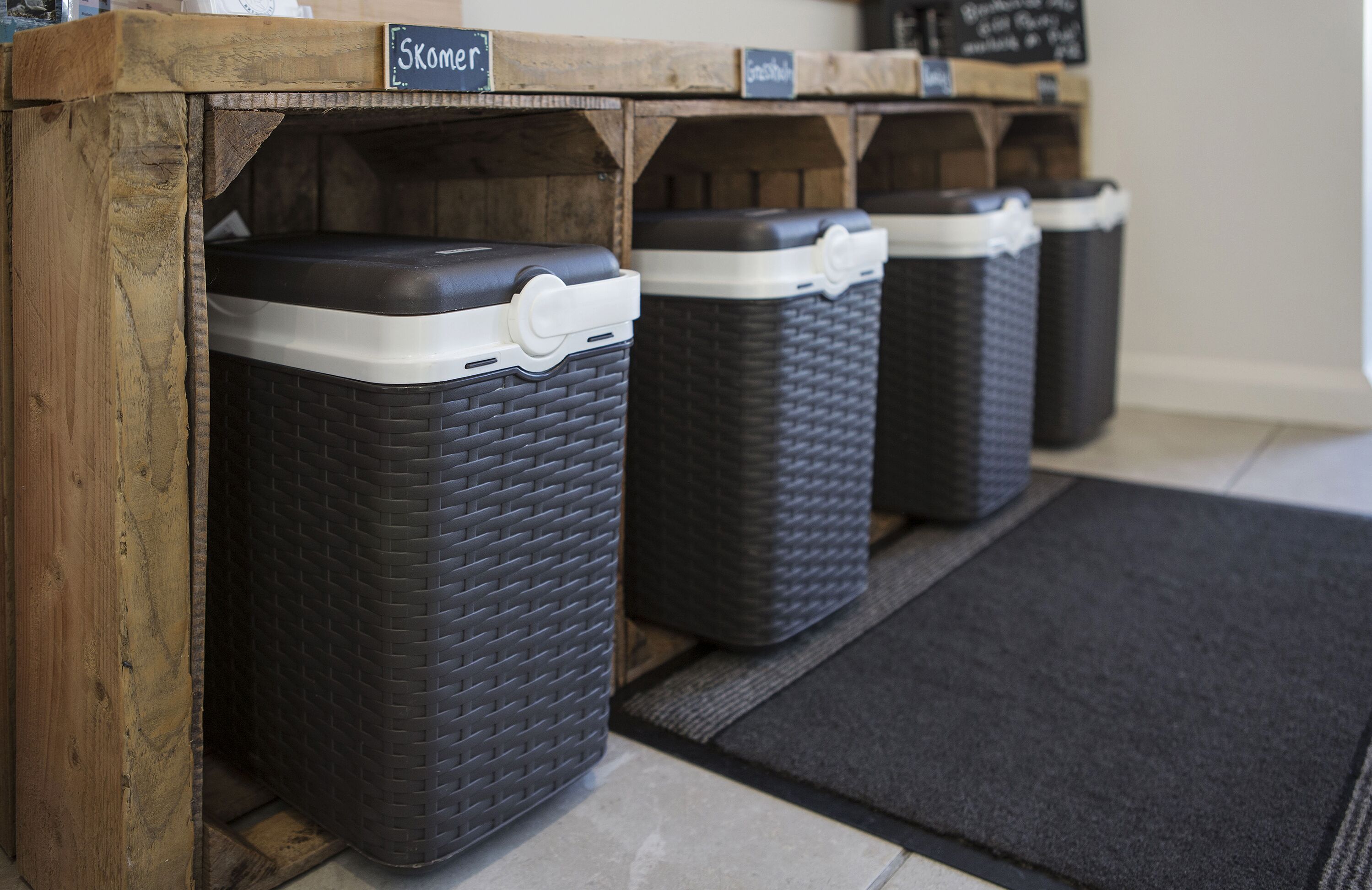 A row of black trash bins with white lids, labeled 'Skomer', under a wooden shelf.