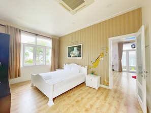A bedroom with a single bed, a nightstand, a wall-mounted lamp, a window with curtains, and a door leading to another room. - Sealink Villas Phan Thiet  Mui Ne (Phu Thuy)