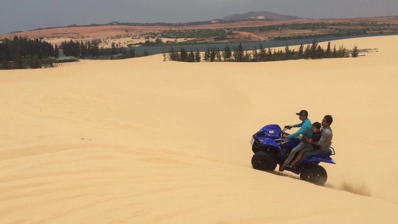 Eine Gruppe von Personen in einem Geländewagen in einer Wüstenlandschaft mit Sanddünen und fernen Hügeln.