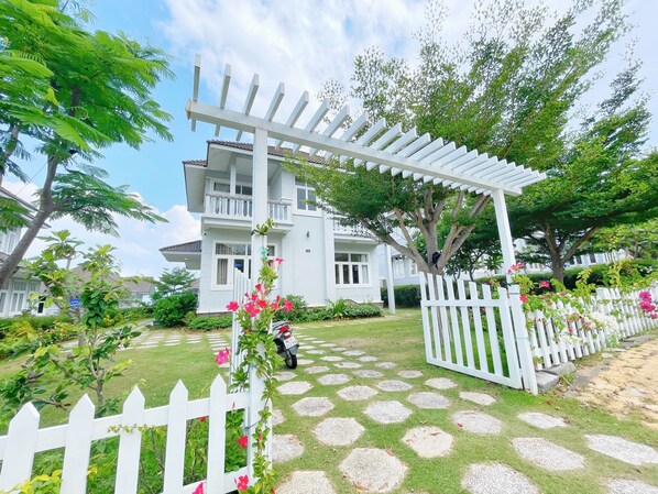 A two-story white house with a balcony, surrounded by a white picket fence and a garden with a patterned stone pathway. - Sealink Villas Phan Thiet Mui Ne (Phan Thiet)