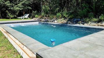 Una piscina rectangular con borde de piedra, rodeada de césped y árboles.