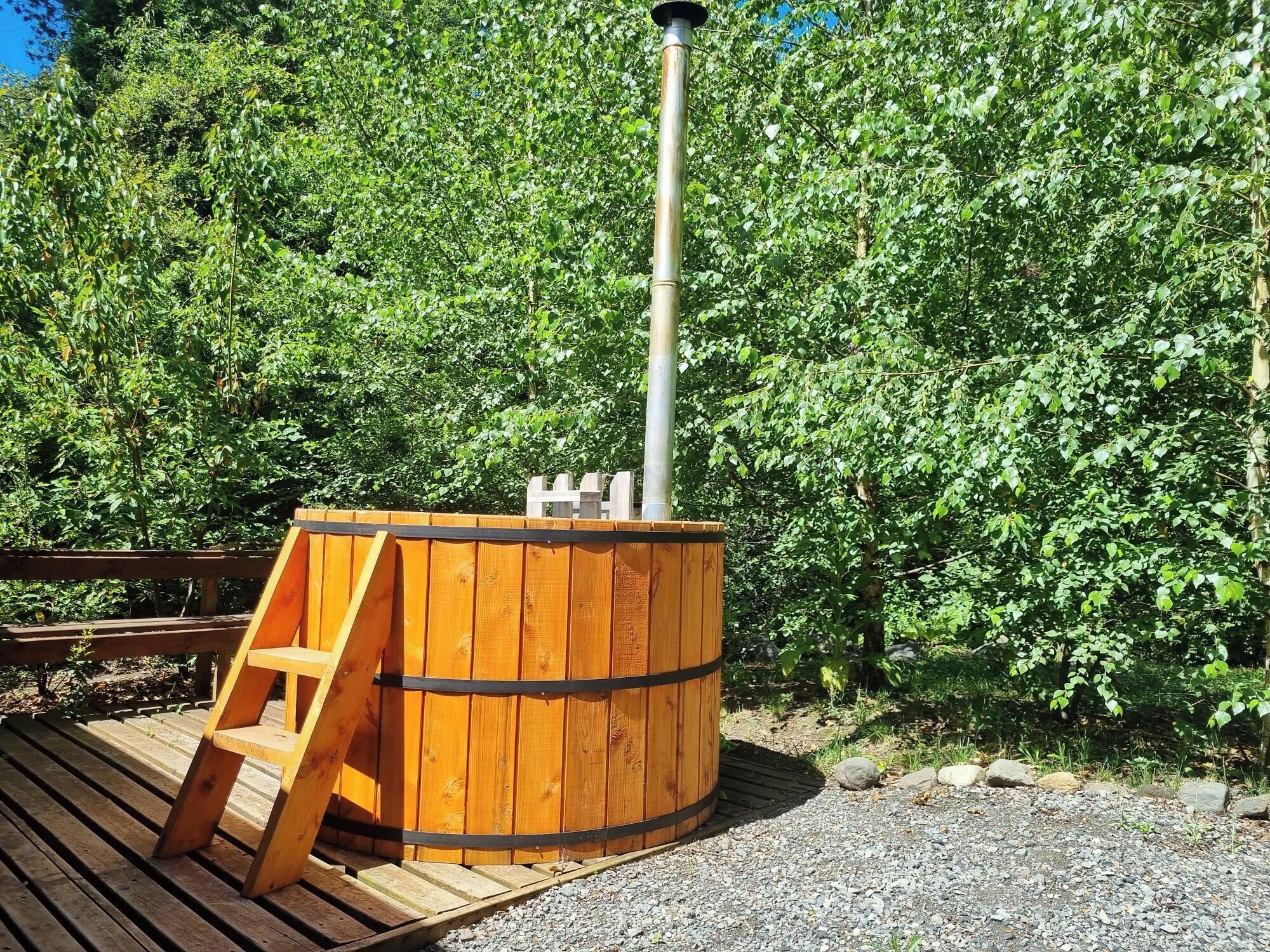 A wooden hot tub with a ladder, set on a wooden deck, surrounded by greenery.