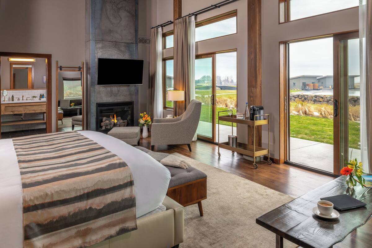 A spacious bedroom with a large bed, a seating area with a chair and table, a fireplace, and a view of the outdoors through large windows.