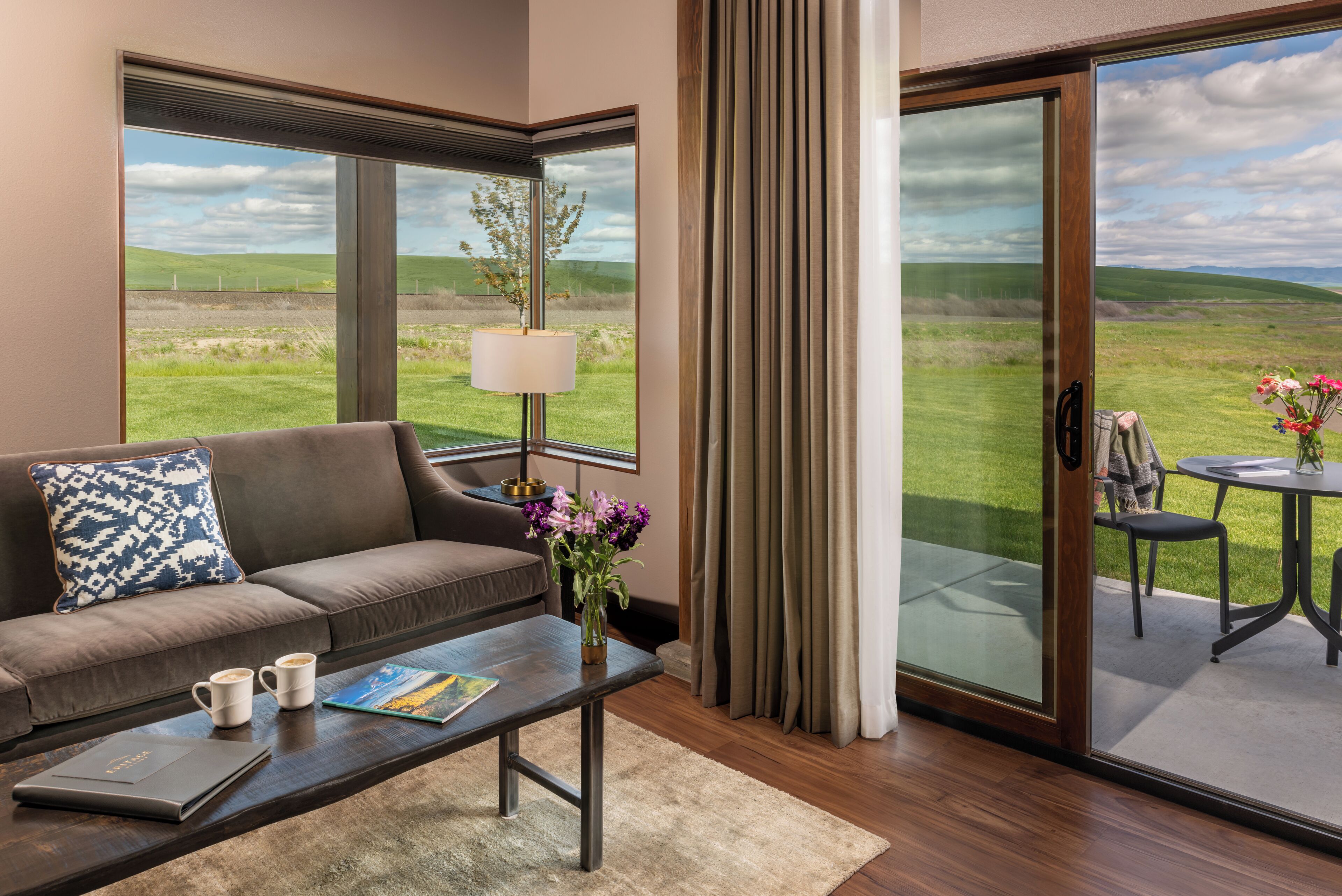 A modern living room with a sofa, coffee table, and a view of a grassy landscape through large windows.