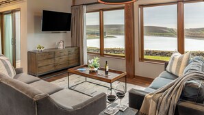 Una sala de estar moderna con un sofá, una mesa de centro, un televisor y ventanas amplias que ofrecen vistas a un lago y colinas.