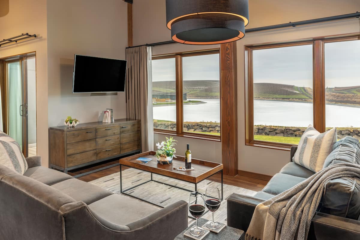 A modern living room with a sofa, a coffee table, a TV, and large windows offering a view of a lake and hills.