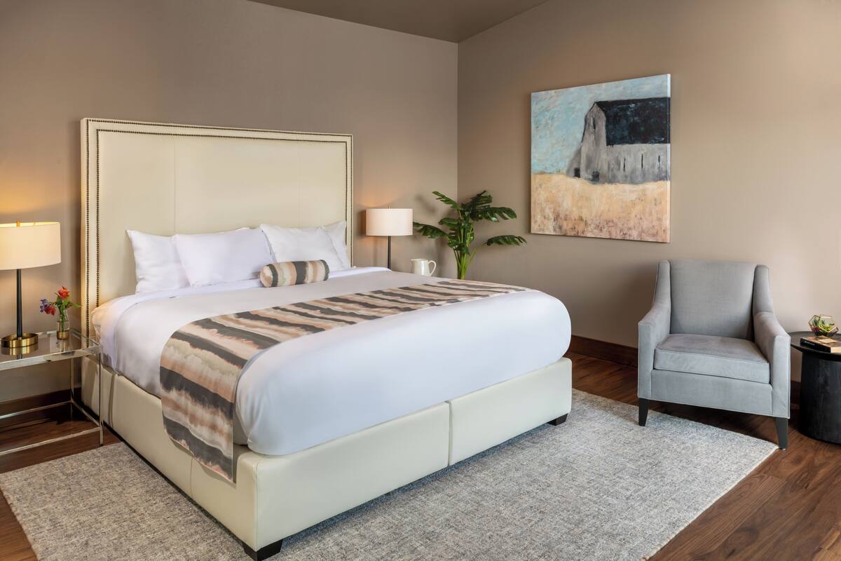 A hotel room with a large bed, a nightstand with a lamp, a chair, and a painting on the wall.
