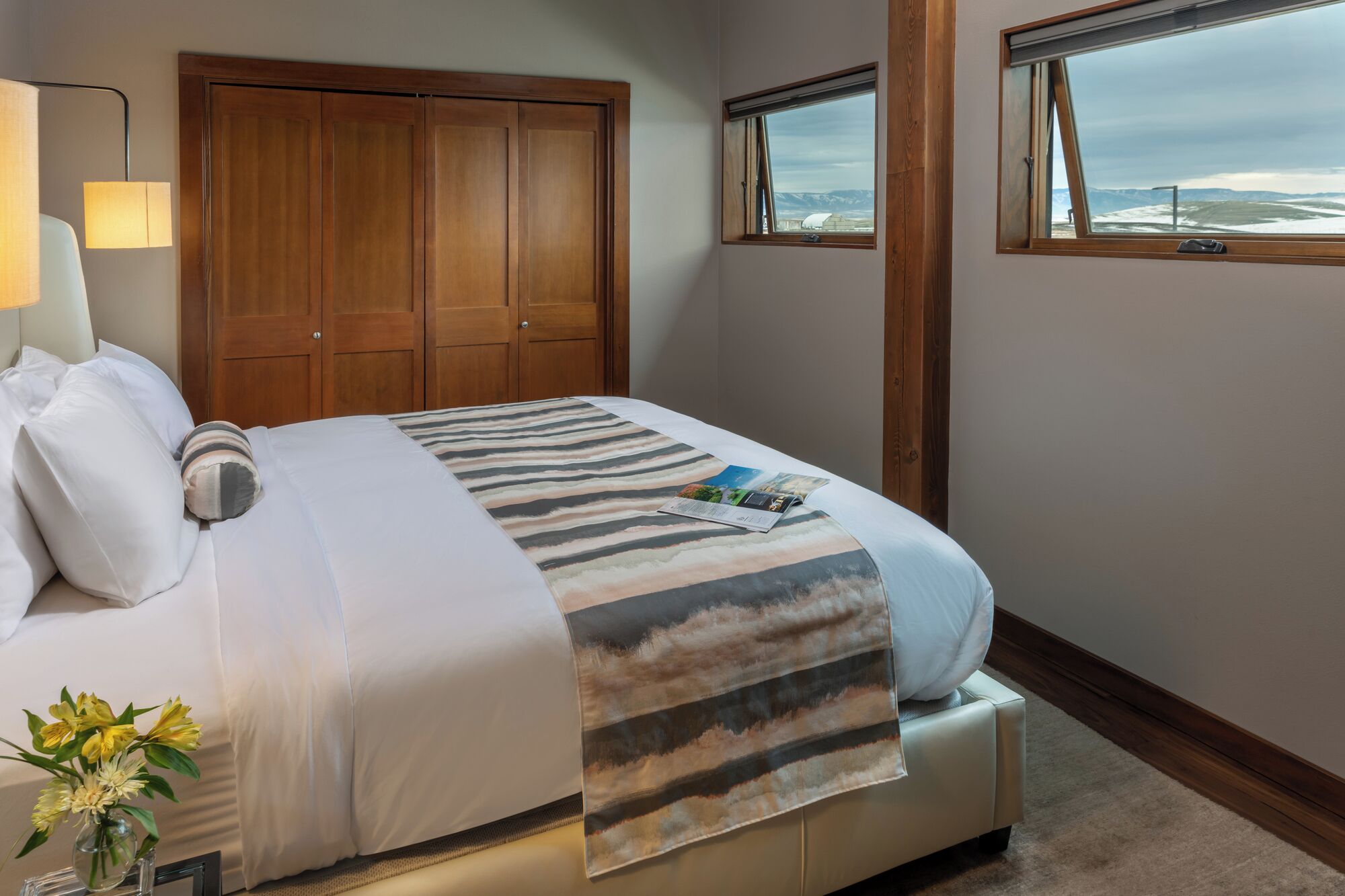 A neatly made bed with a striped blanket, a bedside lamp, and a window with a view of mountains.