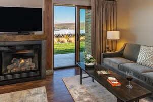 A modern living room with a fireplace, a sofa, a coffee table with books and a glass of wine, and a view of a lake through a sliding glass door.