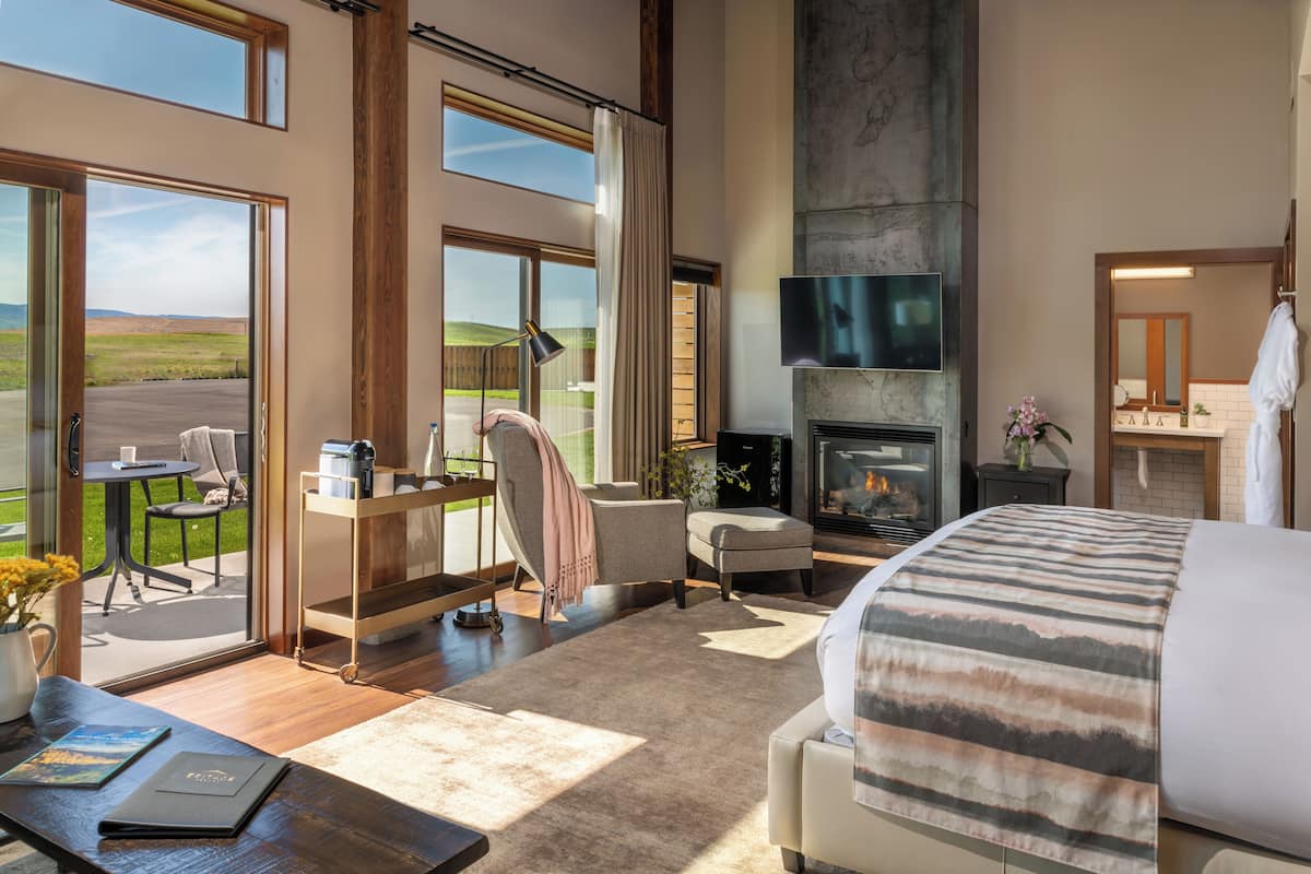A spacious bedroom with a large bed, a fireplace, a desk with a chair, and a view of the outdoors.
