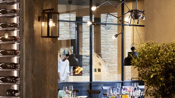Un interior de restaurante con mobiliario de madera, una pared de ladrillo y vista a un cocinero trabajando.