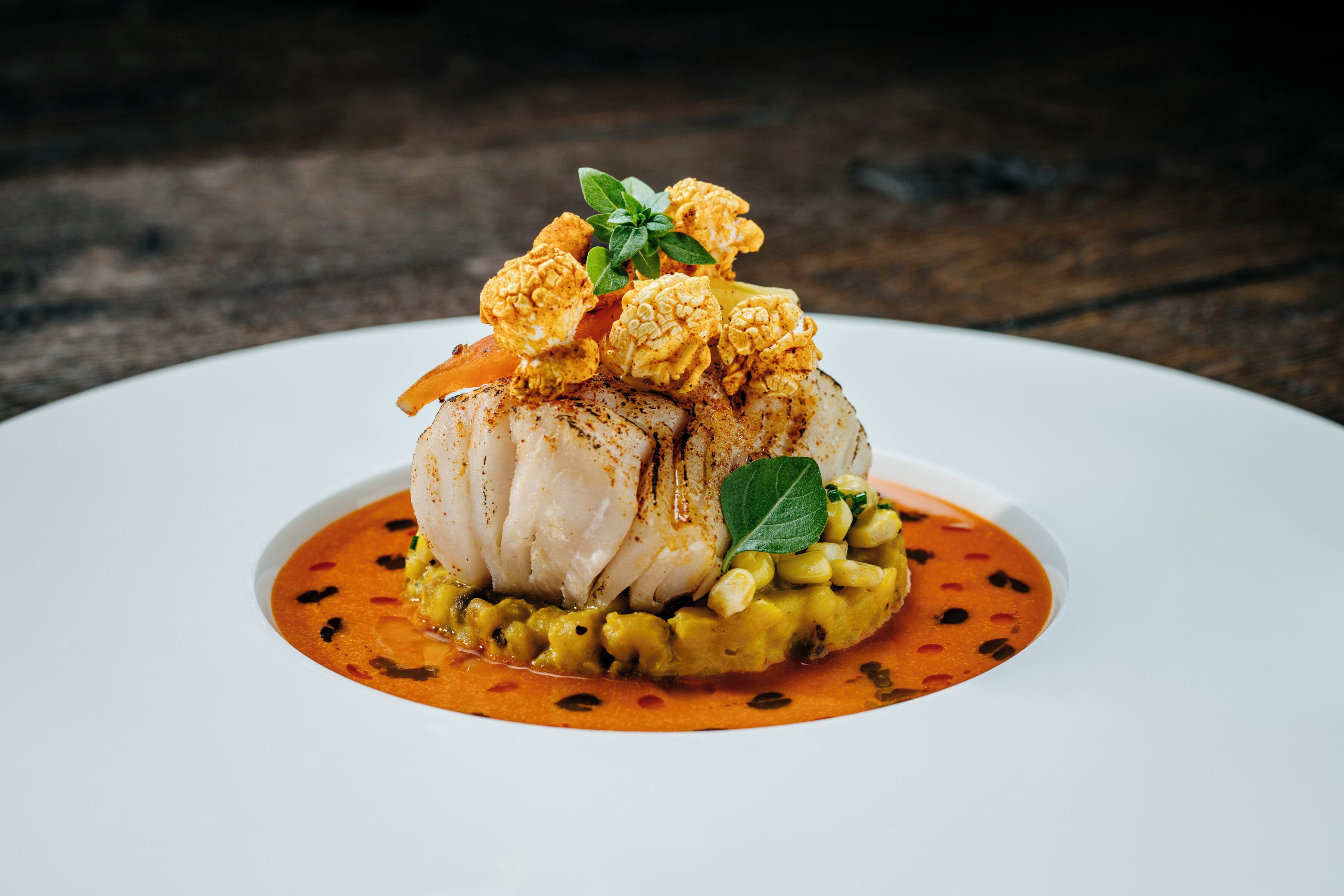 Un plato blanco con un guiso de pescado, adornado con maíz y salsa roja.