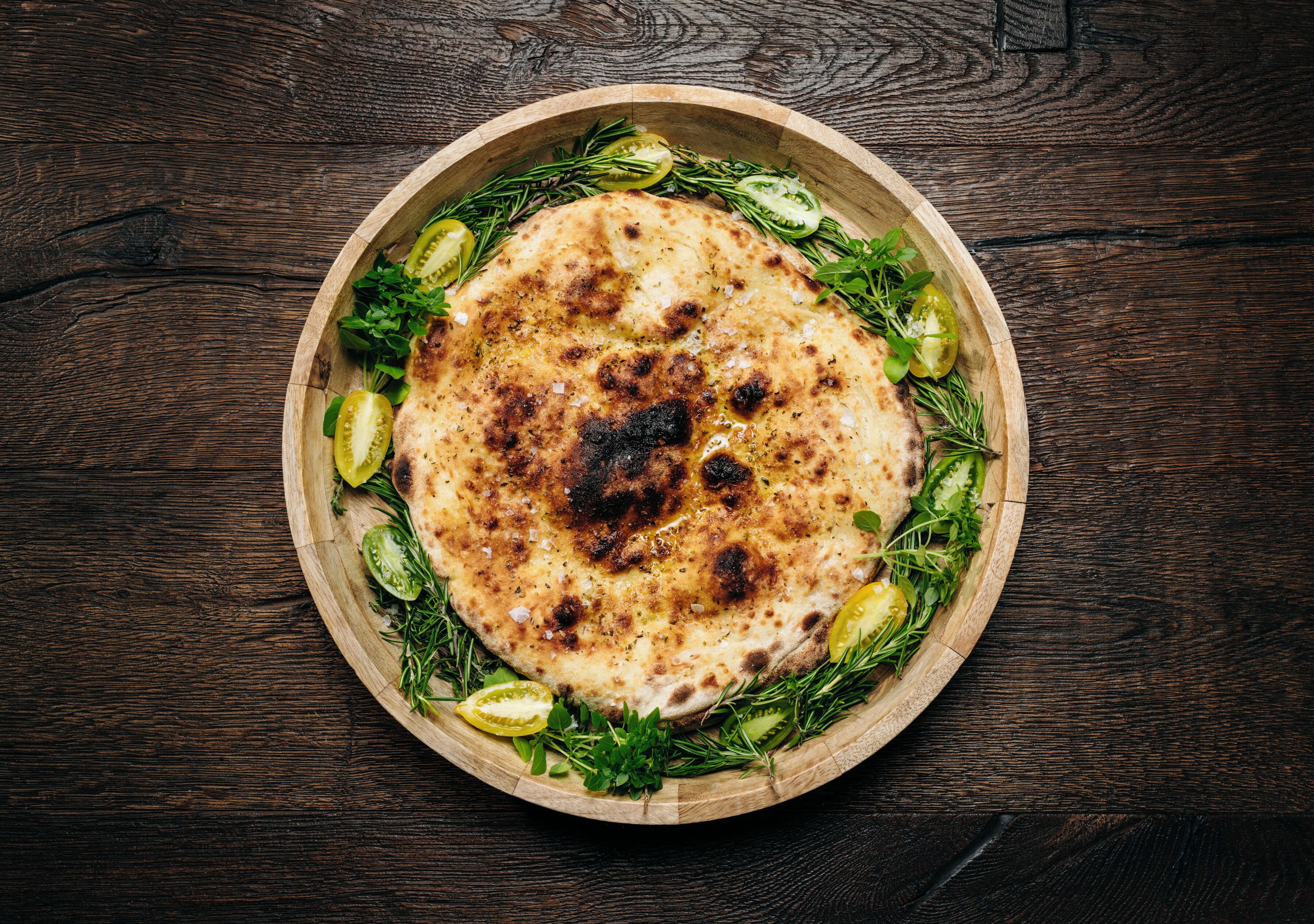 A round wooden board with a flatbread topped with herbs and olive slices.