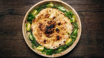 A round wooden board with a flatbread topped with herbs and olive slices.