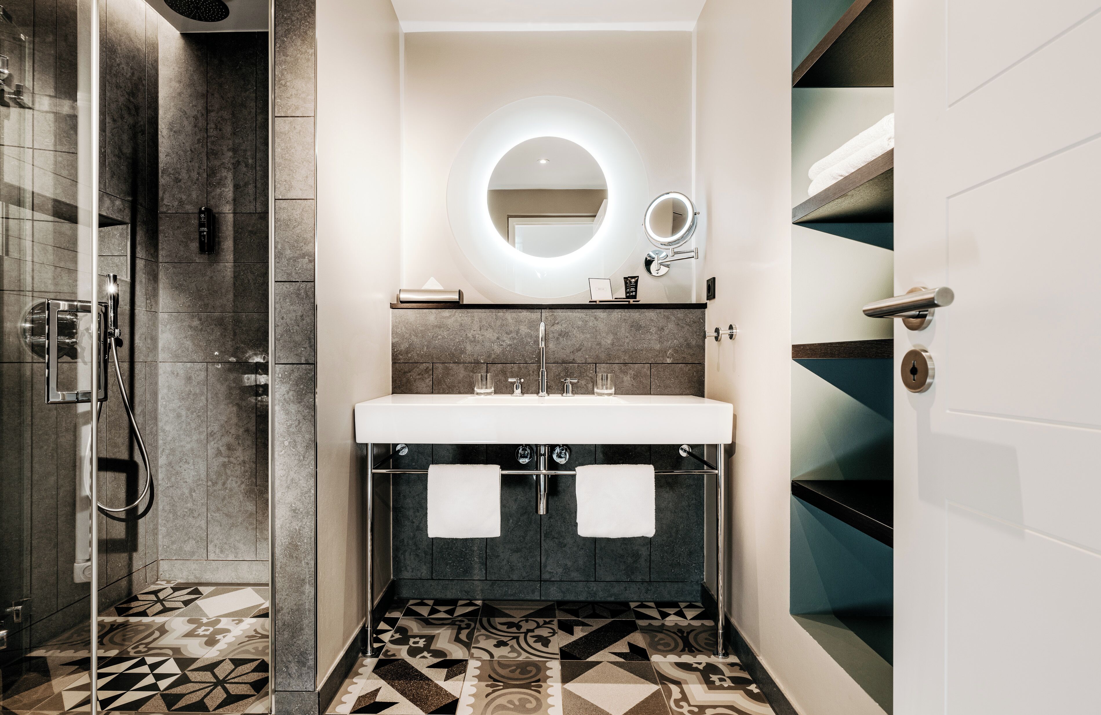 Un baño moderno con ducha, lavamanos, dos toalleros, espejo y suelo estampado.